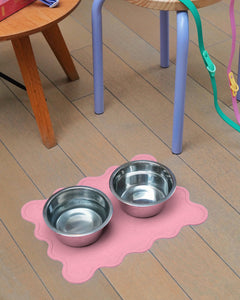 Two metal bowls on a pink mat with a small stool and dog leashes.